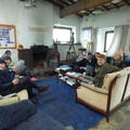 A group of white people sit in a rustic common room on sofas. Many are wearing warm clothing, and some are holding mugs. All are looking towards the camera and smiling