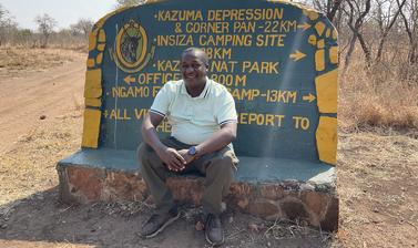 Godfrey Mtare is sitting in front of a sign with spiky shrubs and trees in the background