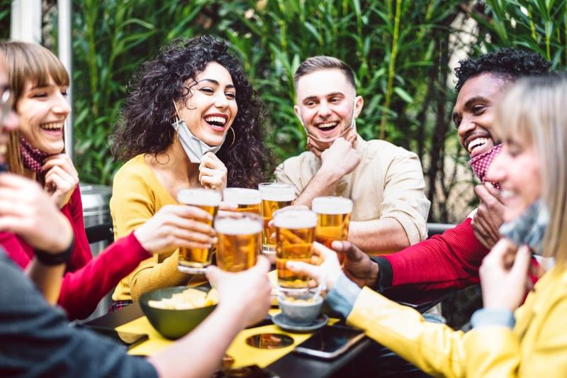 A group of people smile and cheers glasses of beer. All are lowering their face masks