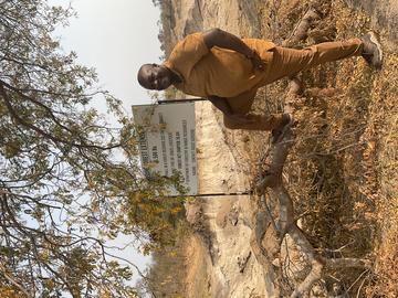 Godfrey Mtare has his foot on a bank of mud, with trees and a sign visible behind. He is looking toward the camera and smiling