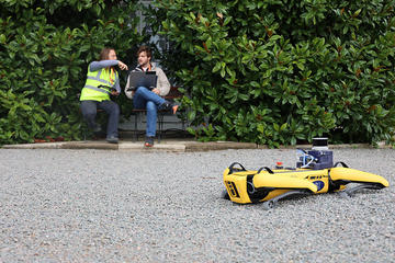 A robot dog rests on gravel in the foreground, tired from its alienated labours. In the background, two humans wearing yellow high-vis jackets pretend to have a conversation. What are they discussing? Maybe cybersynthetic conscioussness - perhaps not...