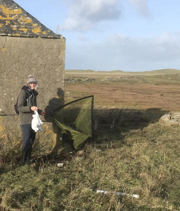 Vallay fieldwork The photo is taken outside. A researcher wearing winter gear is stood next to a grey building. They are looking at the camera and smiling. They are holding a large net.