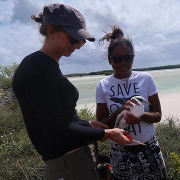 April Burt and a colleague from the Seychelles handle a white-tailed tropicbird. A beach with light blue water is visible in the background.