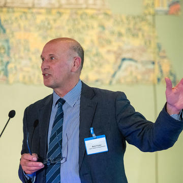 Mark Fricker with his left hand up delivers a speech at a lectern