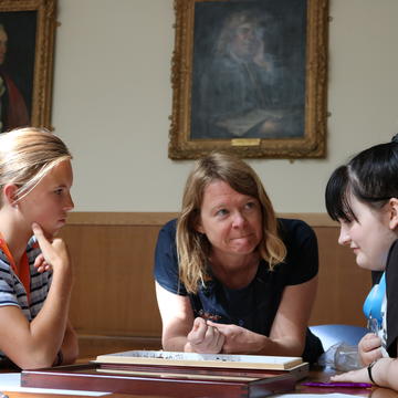 The photo is taken inside. Lindsay Turnbull leans over a table, looking to her right at a group of students. The students are looking down at the table, appearing to speak to each other. A student on the left of the photo is thinking.