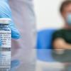 A gloved hand holding a vial of vaccine, with someone sitting out of focus in the background waiting to be vaccinated